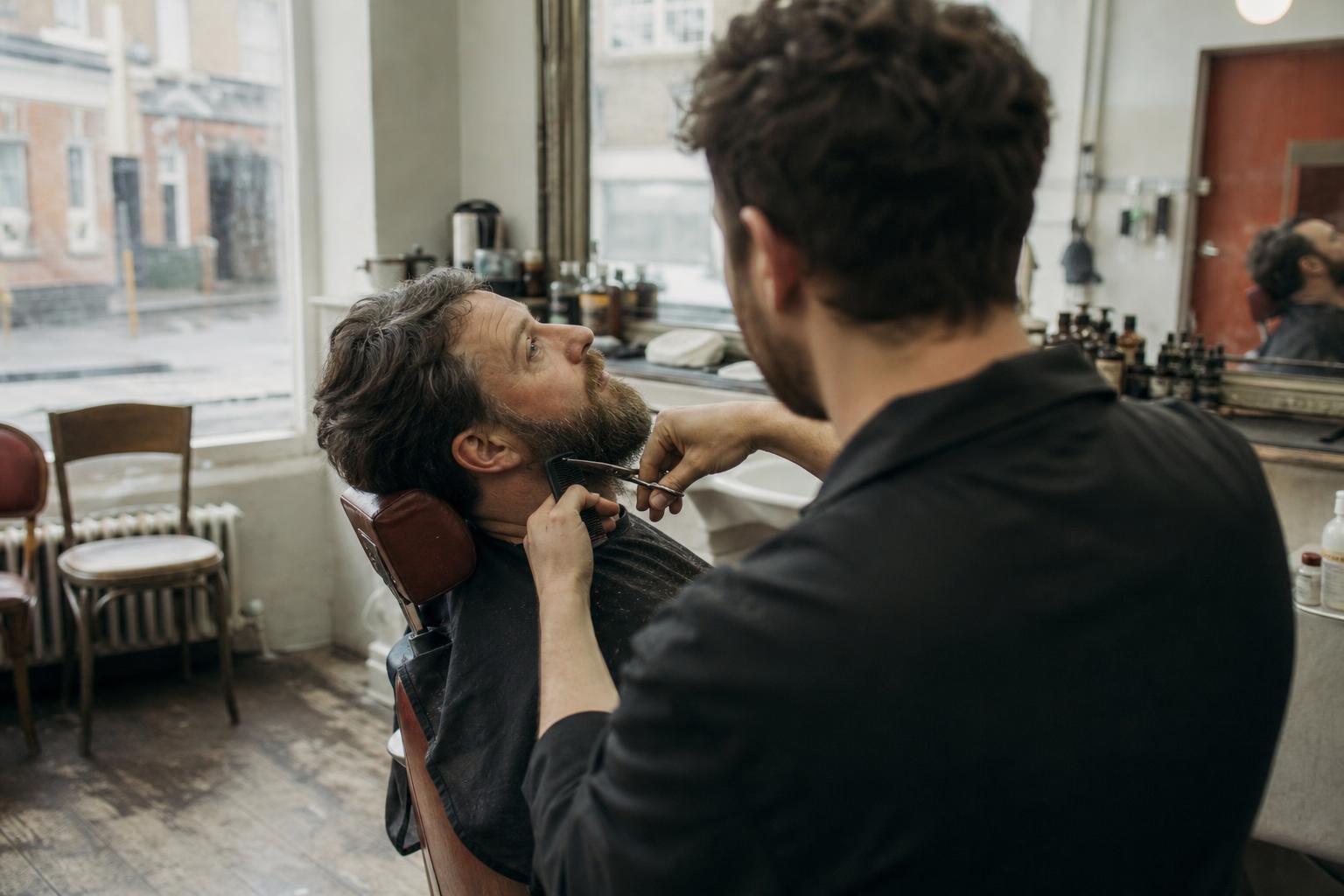 A barber shaping a regular's beard, the booked-in work that competing with a cheap barber is really about