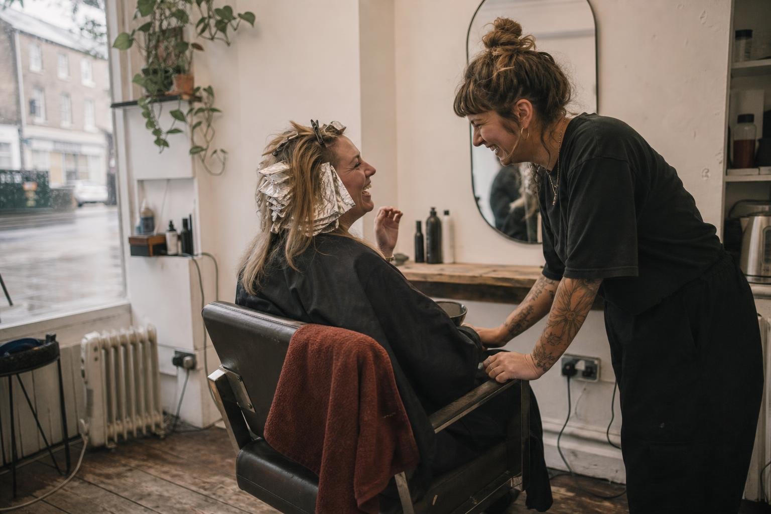A regular client laughing with her stylist mid-colour, foils in, in a small salon