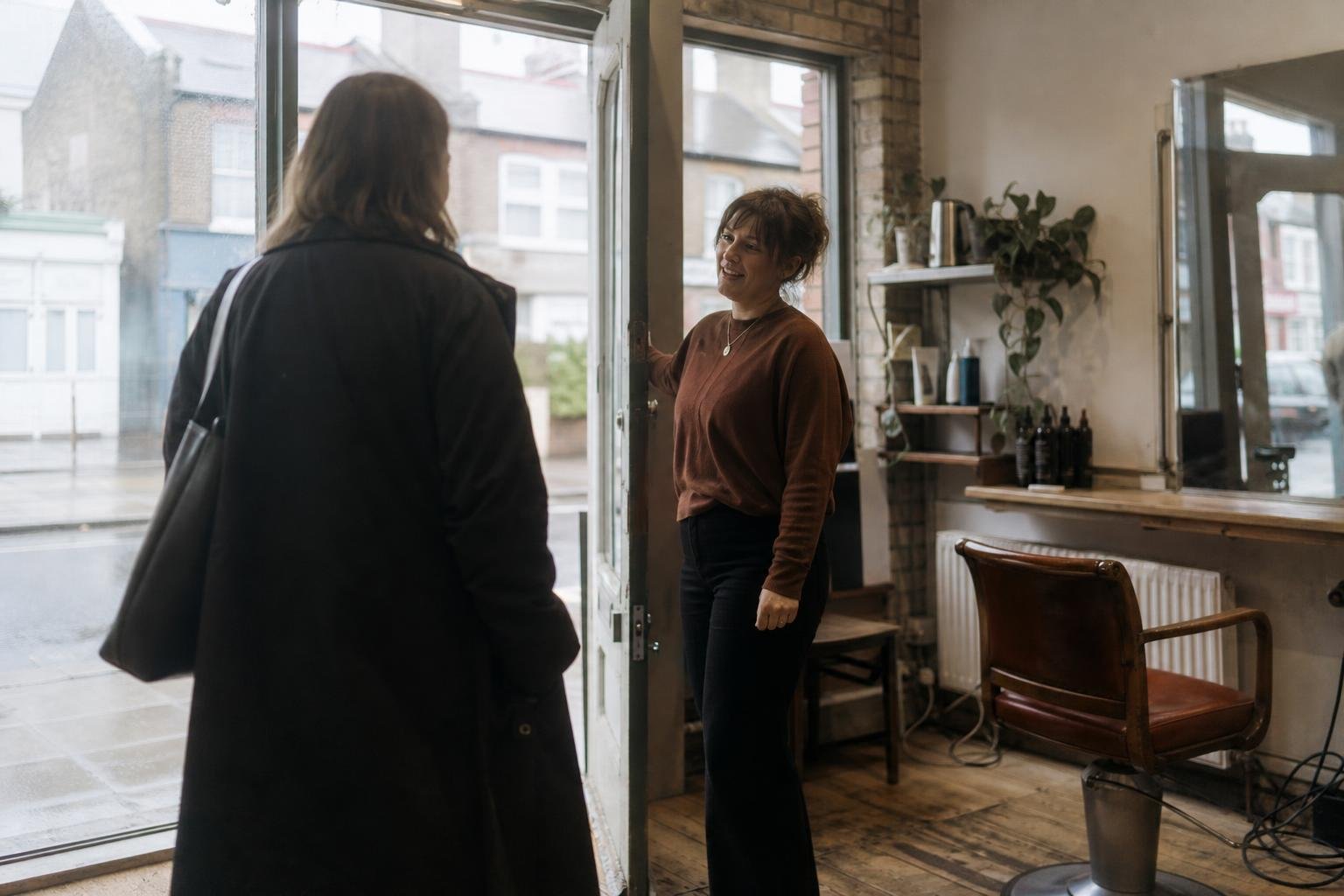 A stylist welcoming a new client at the door of a quiet independent salon, the kind of new client advertising has to pay for