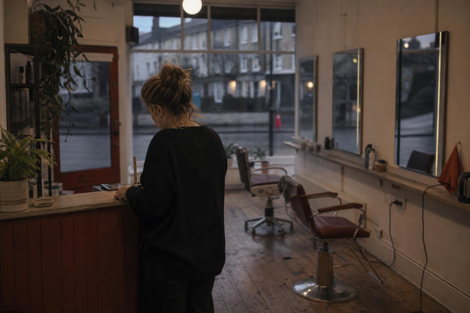 A salon owner alone at her reception counter at closing time, the chairs behind her empty and the street dark outside