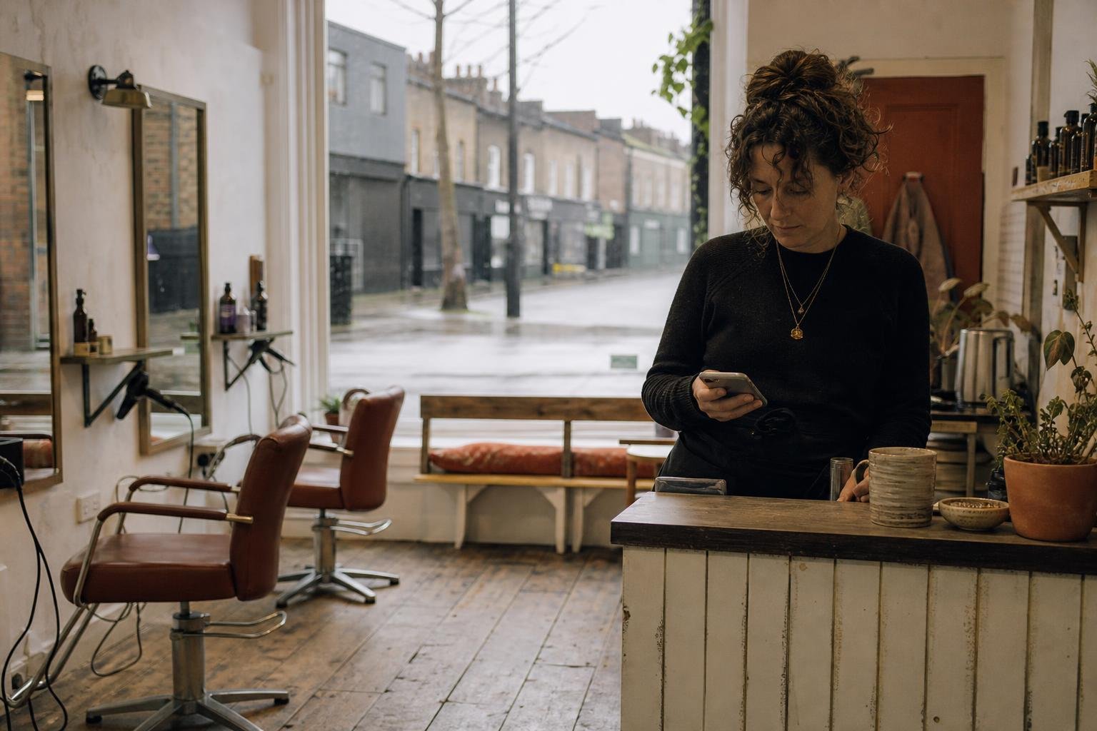 A salon owner checking her phone at the counter beside empty chairs, working out why her salon ads are not showing