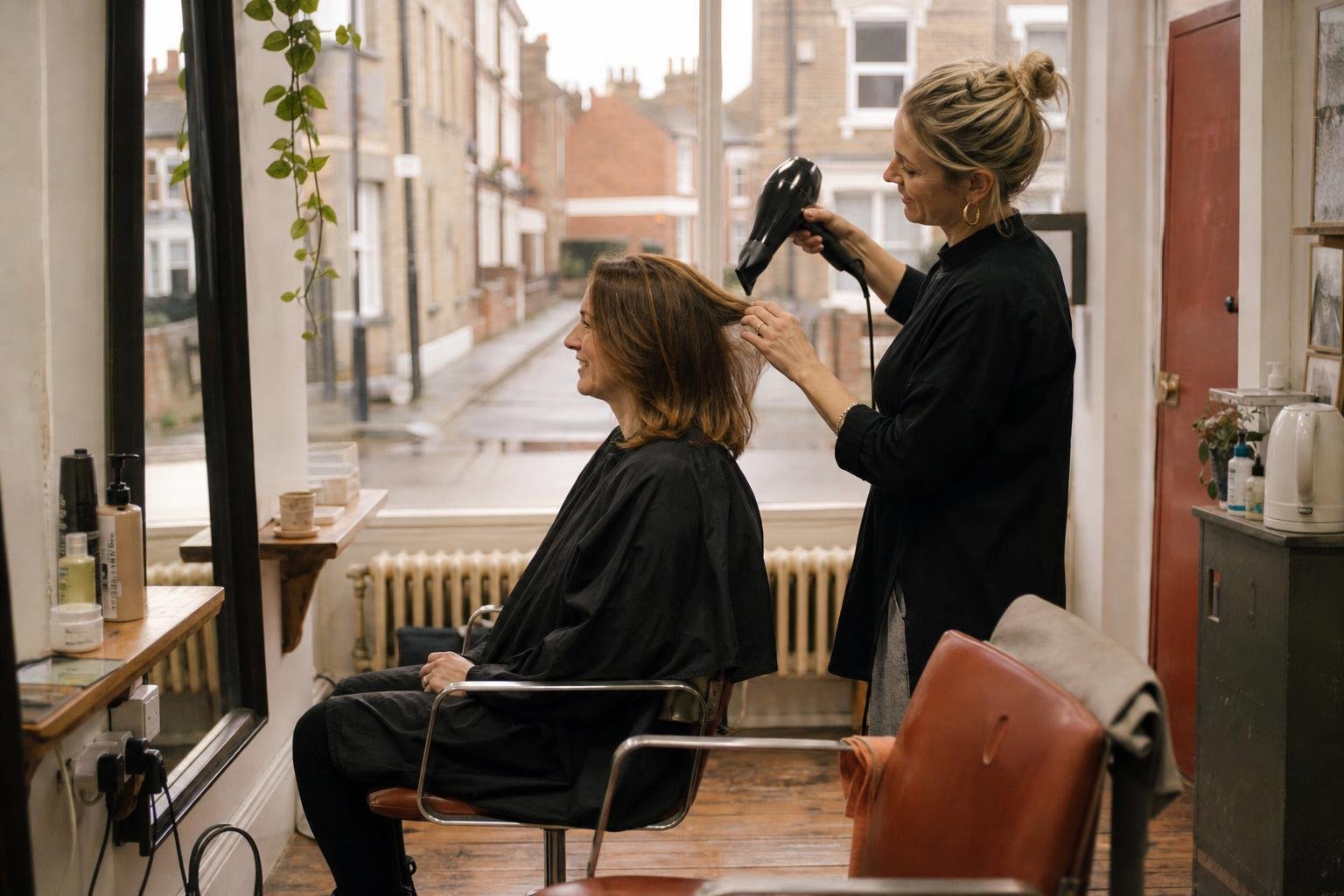 A stylist blow-drying a smiling client in a small salon, the kind of business weighing up a booking app website
